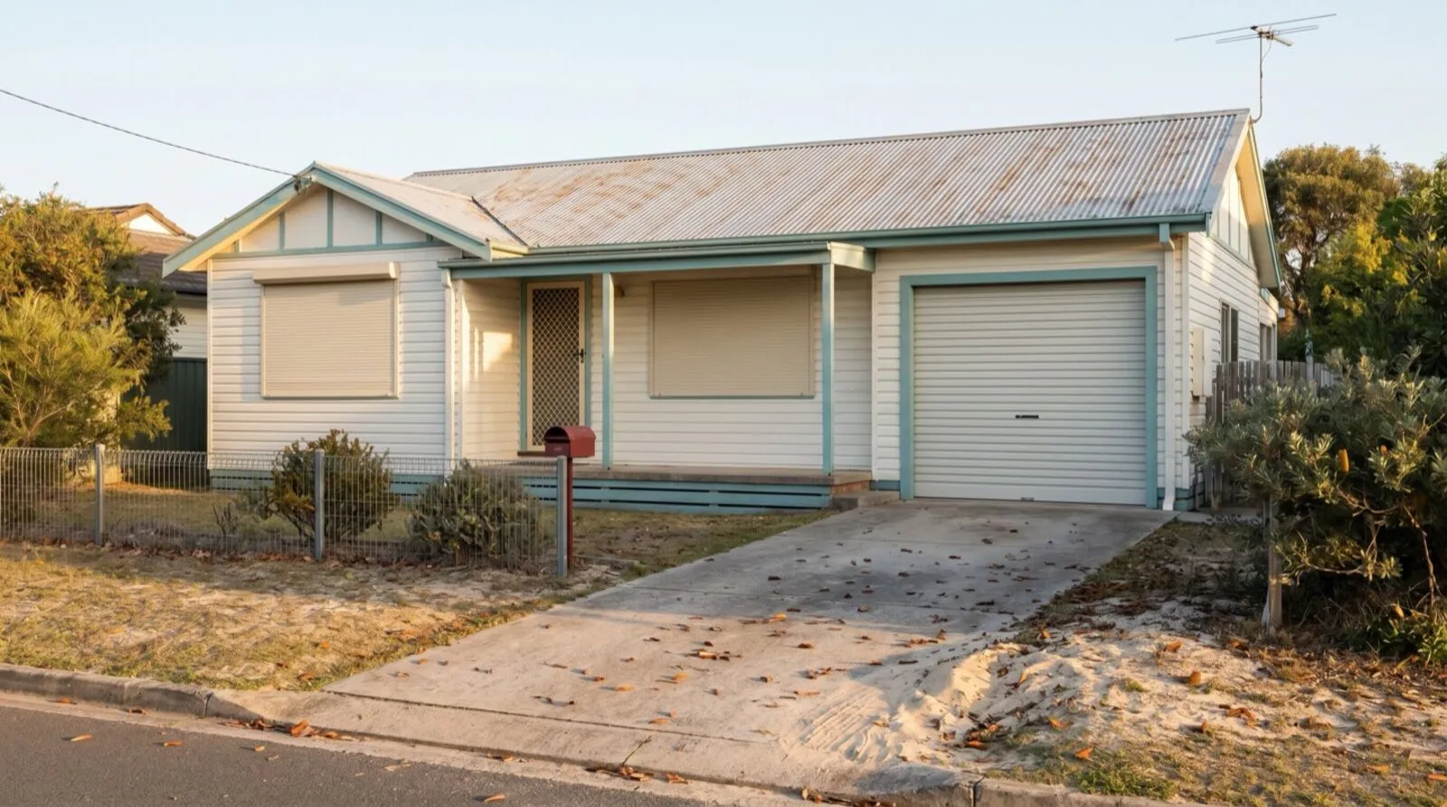 A weatherboard holiday house with a closed roller door, sand and leaves drifted across the empty driveway